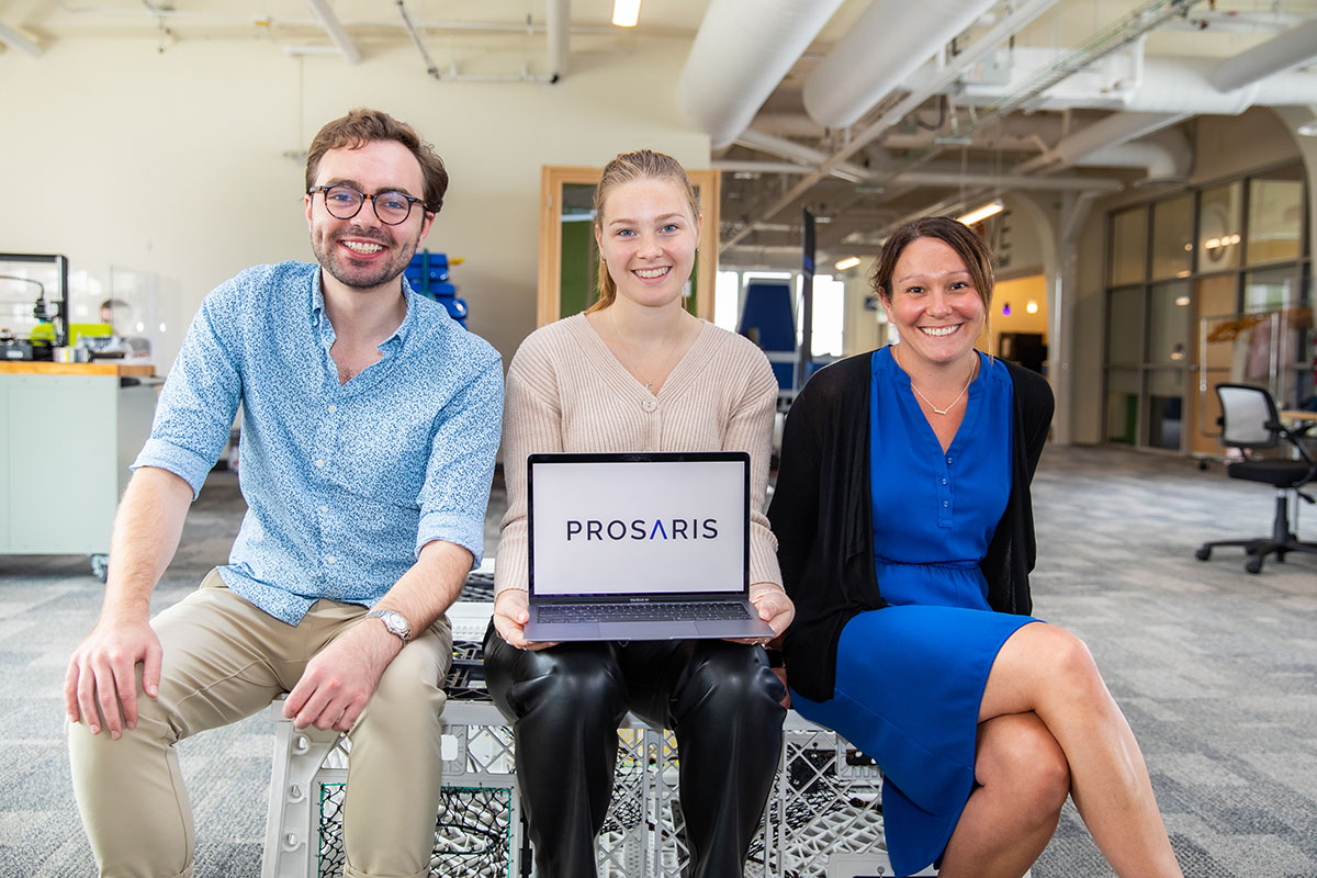 Three people sitting down, the middle person holding a laptop that's open with the Prosaris logo on screen.