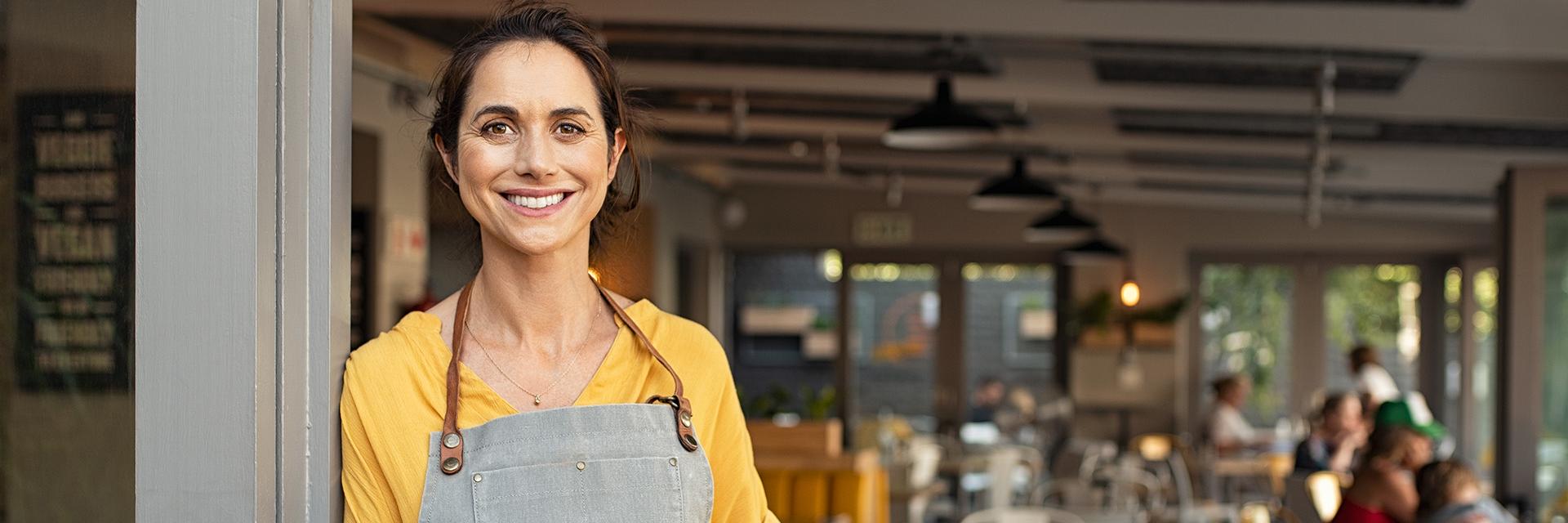 Portrait of happy woman standing at doorway of her store holding digital tablet. Cheerful mature waitress waiting for clients at coffee shop. Successful small business owner in casual clothing and grey apron standing at entrance and looking at camera.