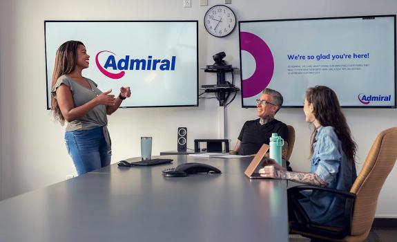 Three people meeting in a conference room. Large screens in the background show the Admiral logo.